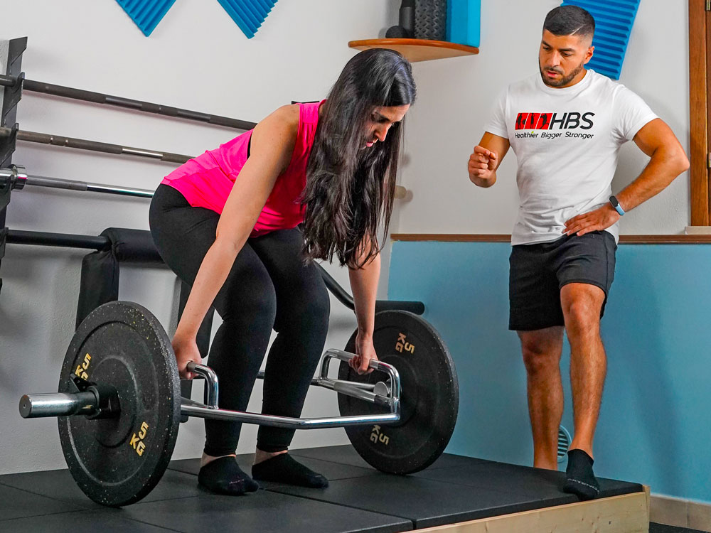 Donna che solleva pesi con bilanciere esagonale, supervisionata da un coach HBS, per valutazione sportiva e nutrizione.