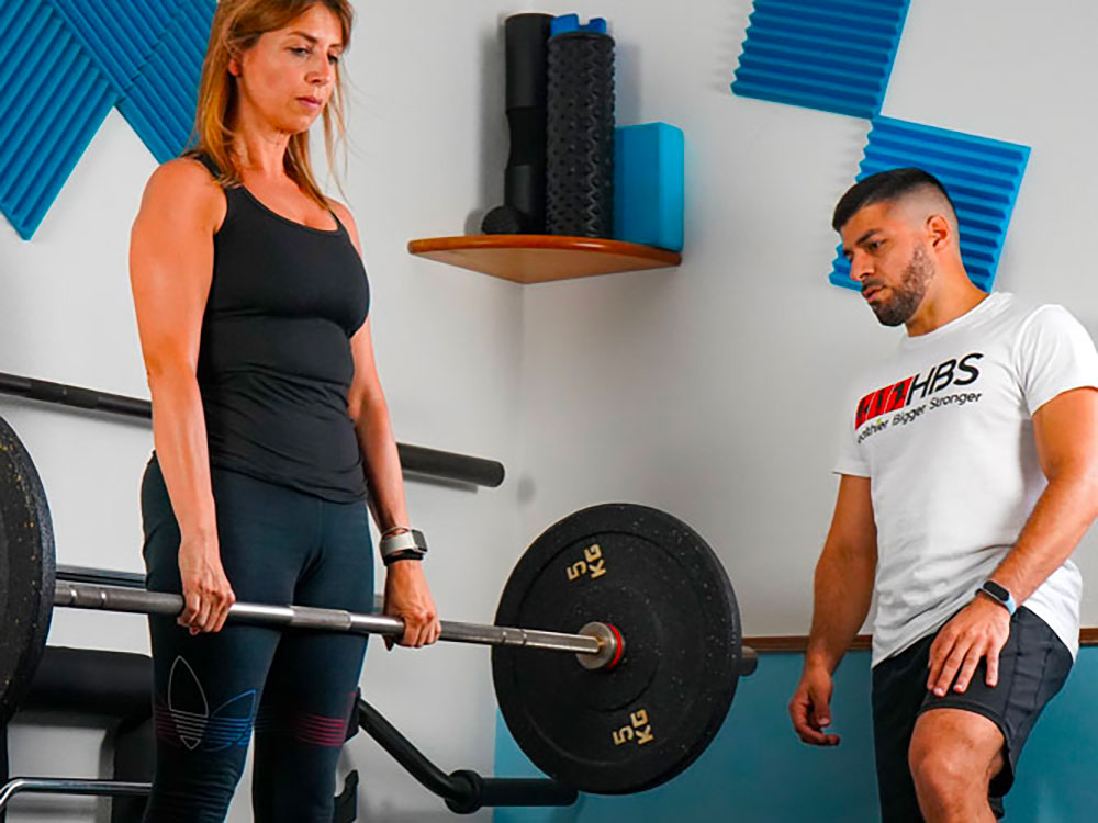 Donna solleva bilanciere con coach HBS, training sportivo per atleti, valutazione atletica e nutrizione.